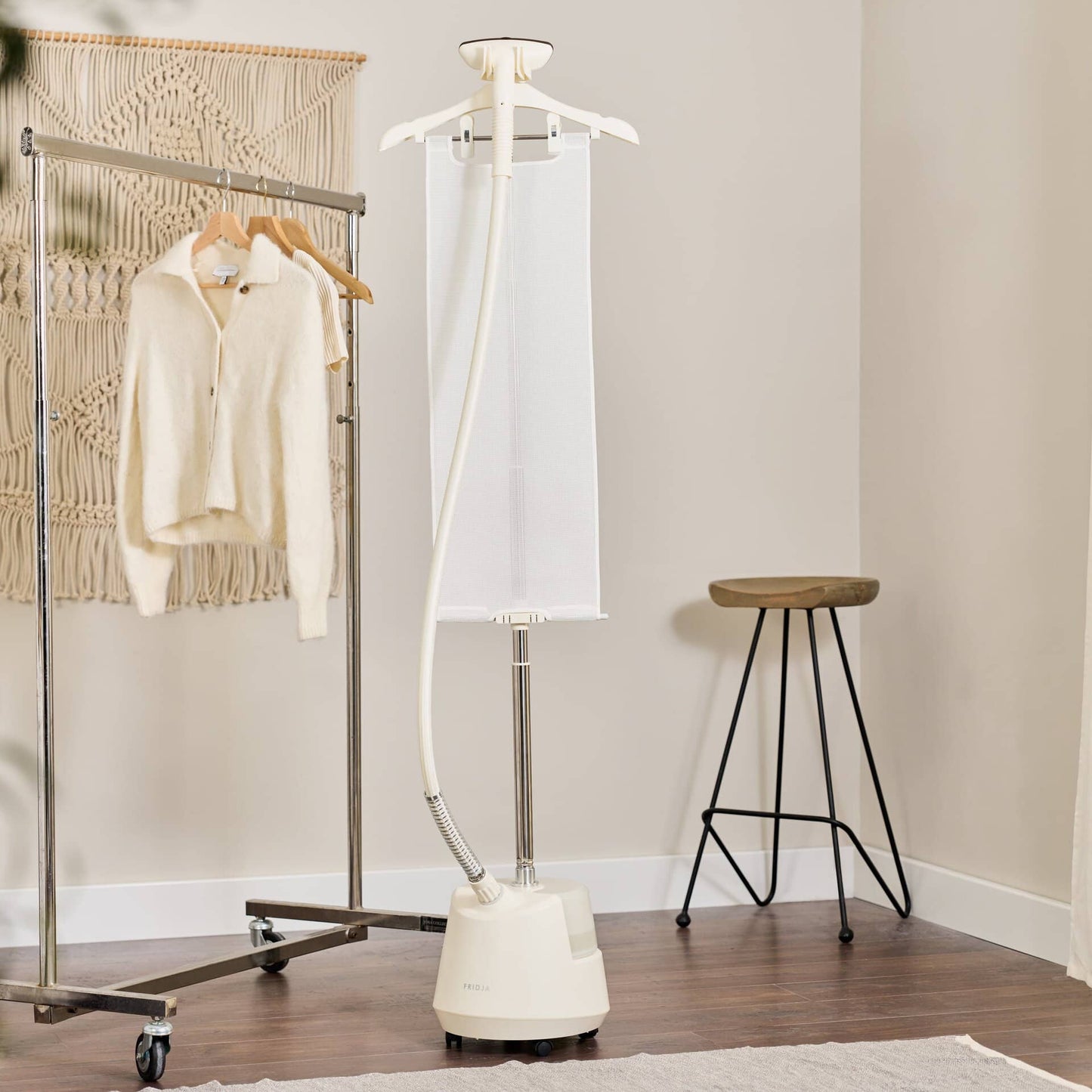 The Fridja f1200 Clothes Steamer Pro stands in a tidy room with a sweater on a clothing rack, macramé wall art, a stool, and wooden flooring.