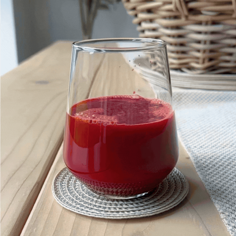 A clear glass filled with red juice sits on a woven coaster atop a light wooden table, with a wicker basket and beige textured cloth in the background.