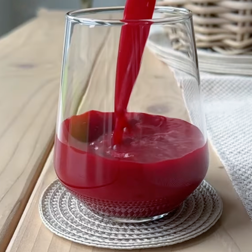 A clear glass is being filled with a thick, bright red juice, sitting on a woven coaster atop a wooden table, with a wicker basket and white cloth in the background.