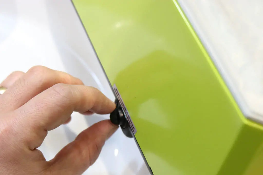 A hand is holding a small suction cup that is attached to a thin pane of glass next to a bright green surface.