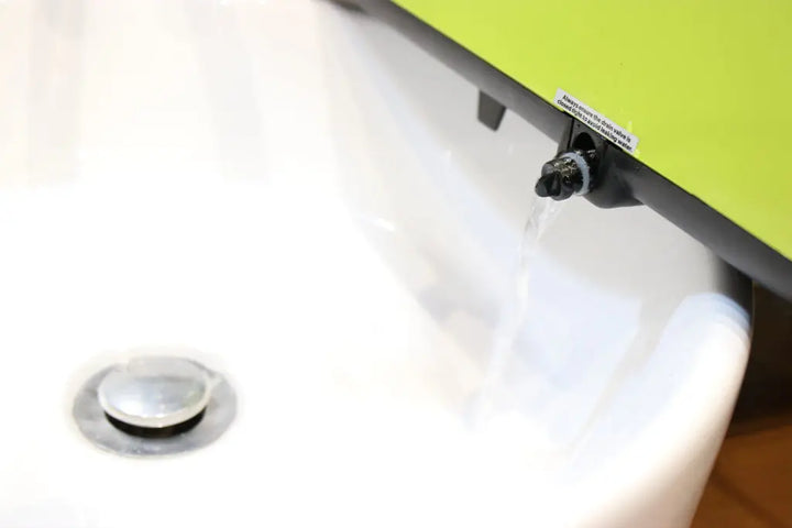 Water trickles from a small faucet on the side of a green object into a white sink, with a silver drain visible at the bottom left.