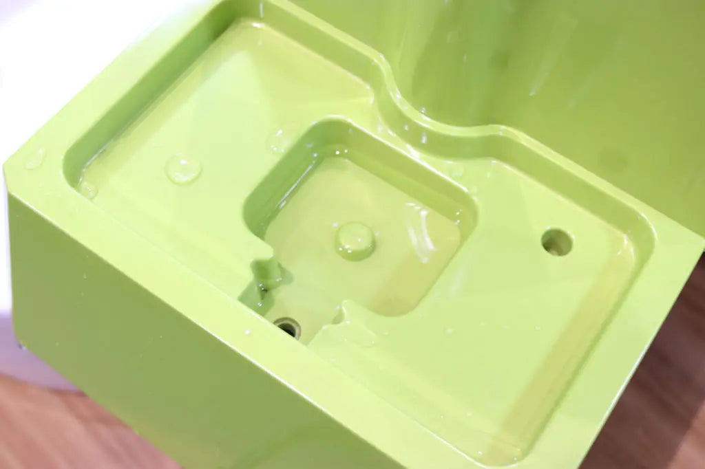 Close-up of a green drinking fountain with water pooled around the drain area, showing the basin’s smooth surface and curved edges.