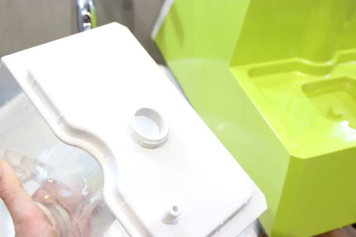 A close-up of a hand holding a white plastic container with a threaded opening, next to a bright green object with molded sections. The background is slightly blurred.