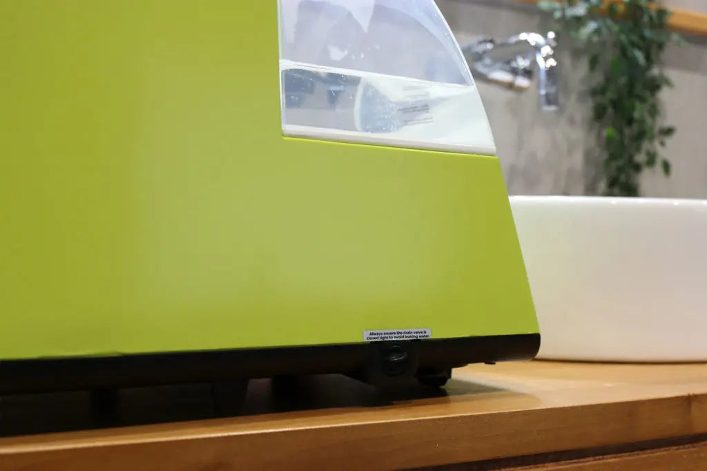 A close-up of a green appliance with a transparent window, sitting on a wooden countertop next to a white sink and faucet, with a blurred leafy plant in the background.