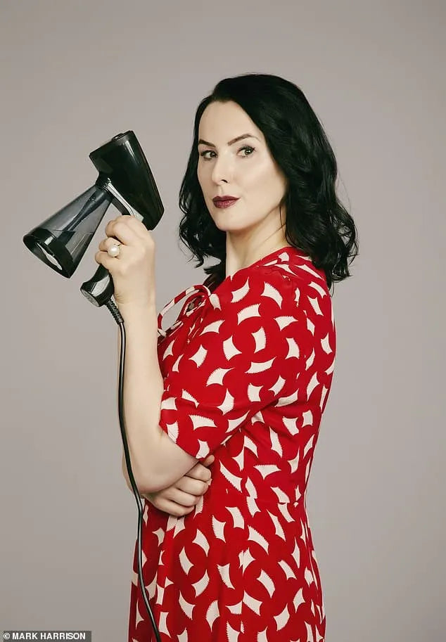 A woman with wavy dark hair wearing a red patterned dress stands against a plain background, holding a steam iron upright and looking confidently at the camera.