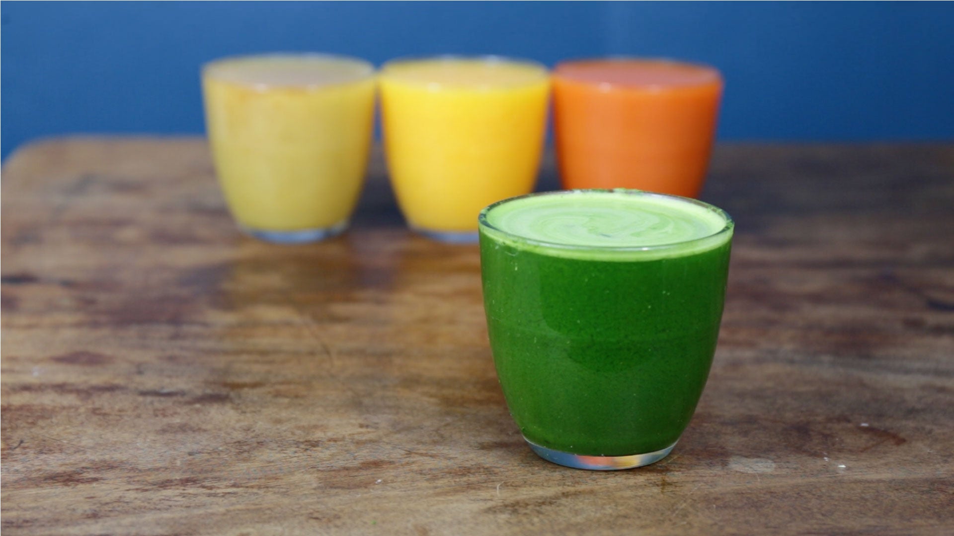 Four glasses of smoothies sit on a wooden surface; the front glass contains a vibrant green smoothie, while the three glasses in the background contain yellow, orange, and pale yellow smoothies.