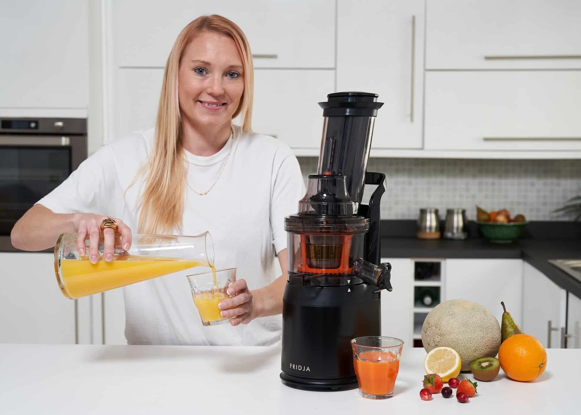 A woman in a white shirt pours orange juice from a glass pitcher into a glass in a modern kitchen. A black juicer, fruits, and two glasses of juice are on the white countertop.