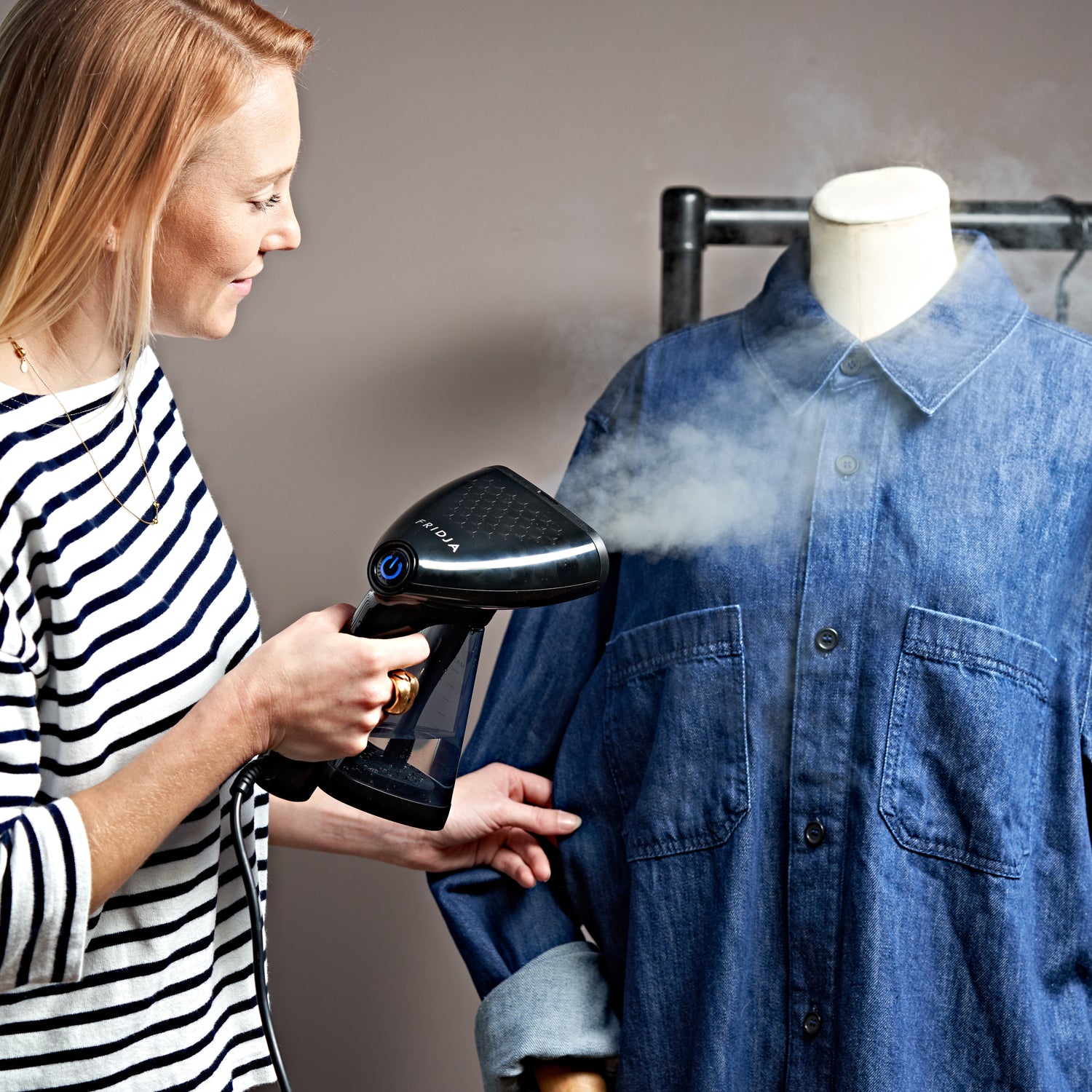 A woman in a striped shirt uses a handheld steamer to remove wrinkles from a blue denim shirt on a mannequin, with steam visible coming from the steamer.