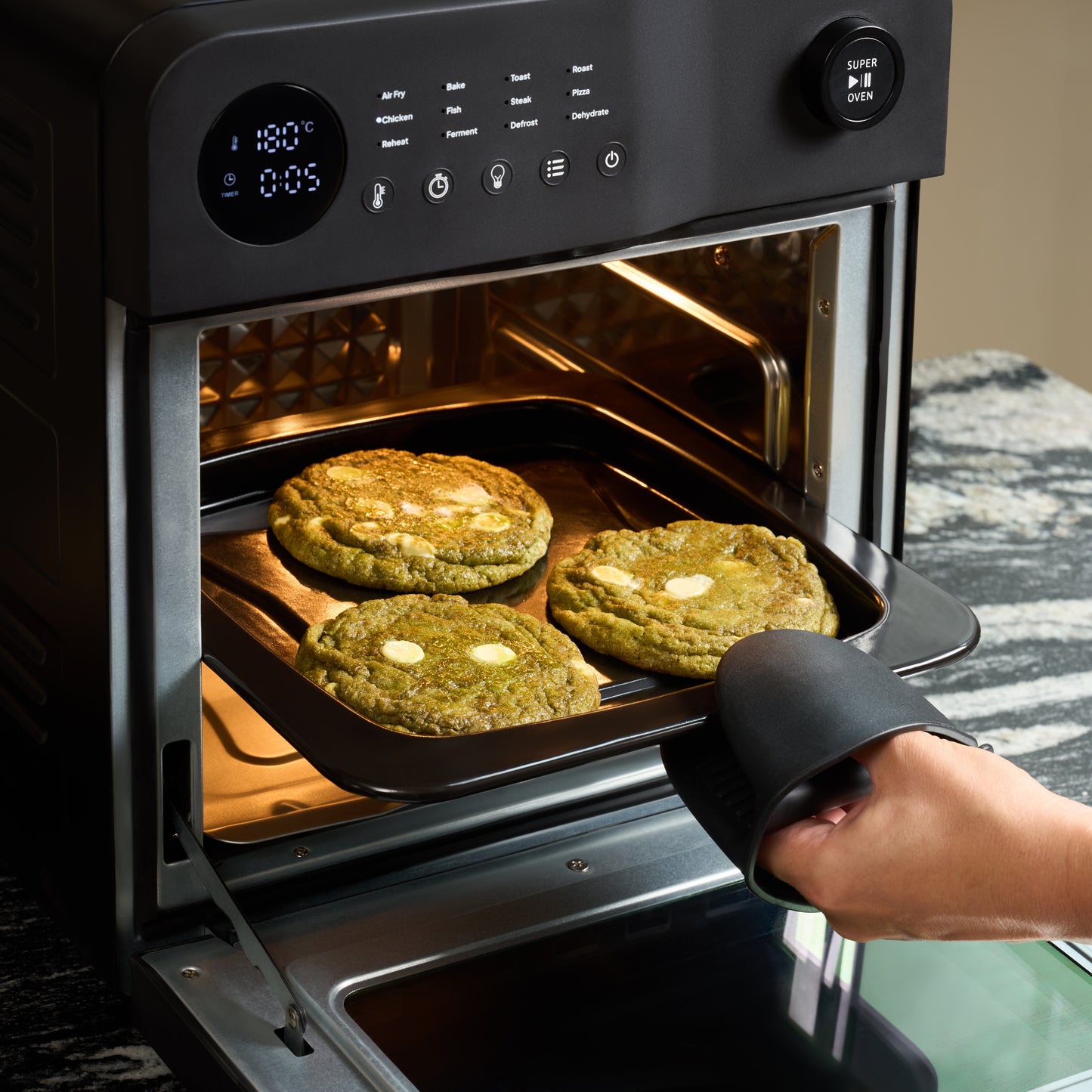 Using a black oven mitt, someone removes a tray with three large green cookies from the Fridja Super Oven - 15L Digital Air Fryer Oven. The convection cooking ensures the white chocolate chip cookies are evenly baked and golden.