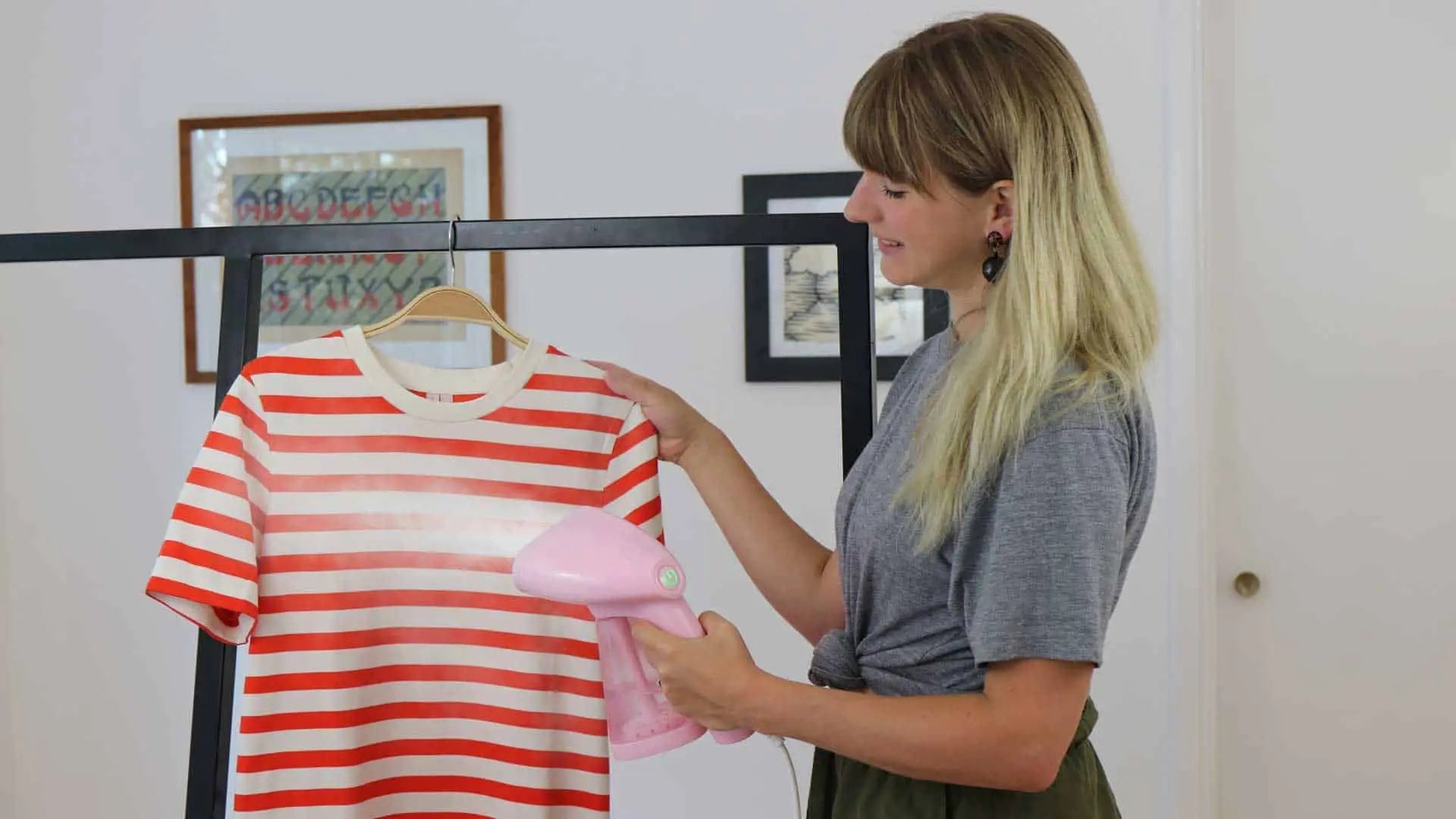 A woman uses a pink handheld steamer to remove wrinkles from a red and white striped T-shirt hanging on a rack in a bright room.