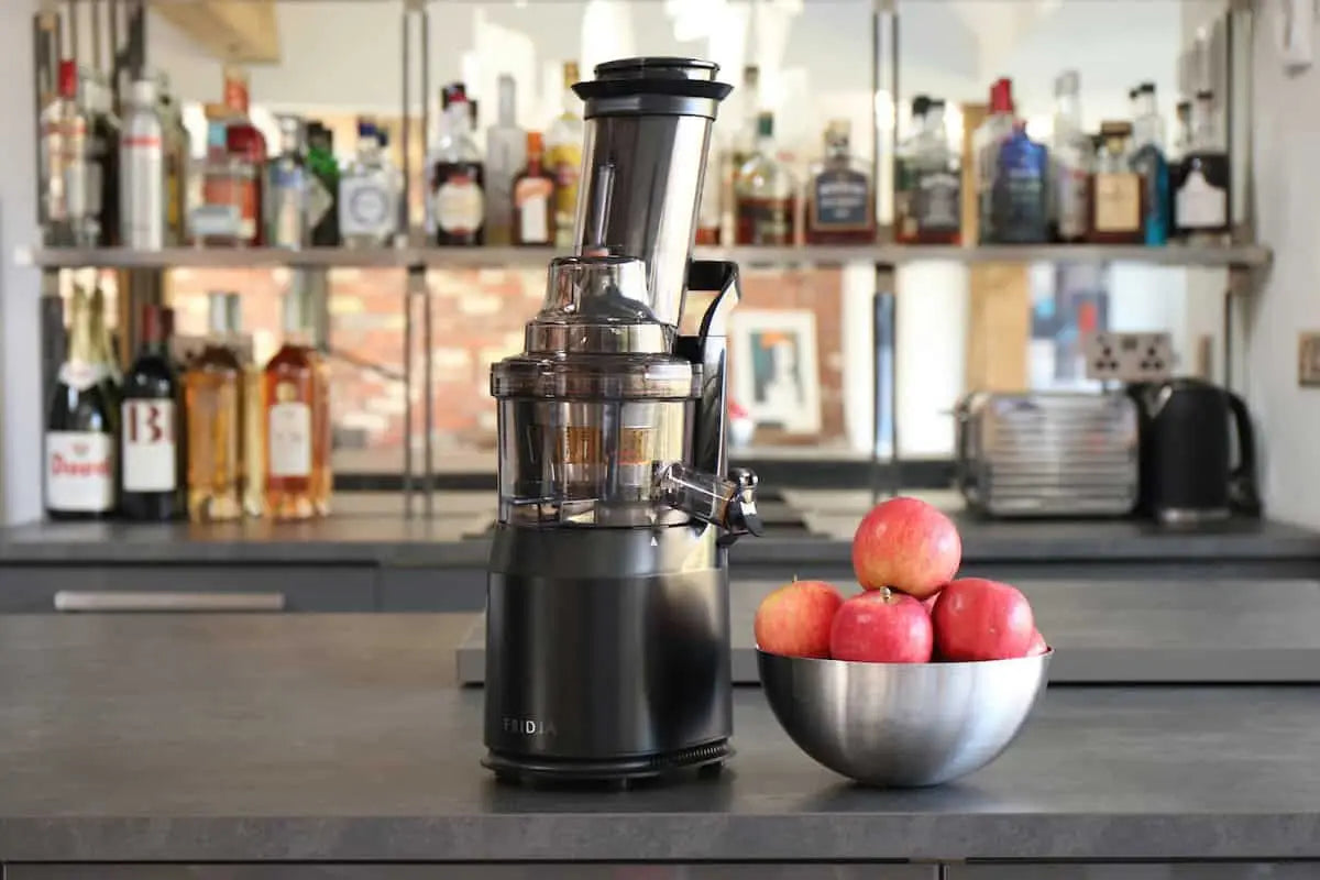 A black juicer sits on a kitchen counter next to a metal bowl filled with red apples. Shelves with assorted bottles and kitchen items are visible in the background.