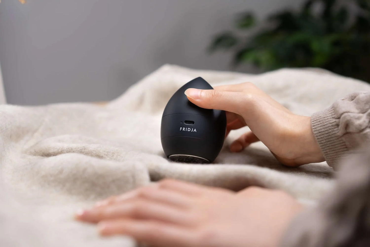 A person holds a small, black, egg-shaped device labeled FRIDJA on a beige fabric surface, with another hand resting nearby. A green plant is blurred in the background.