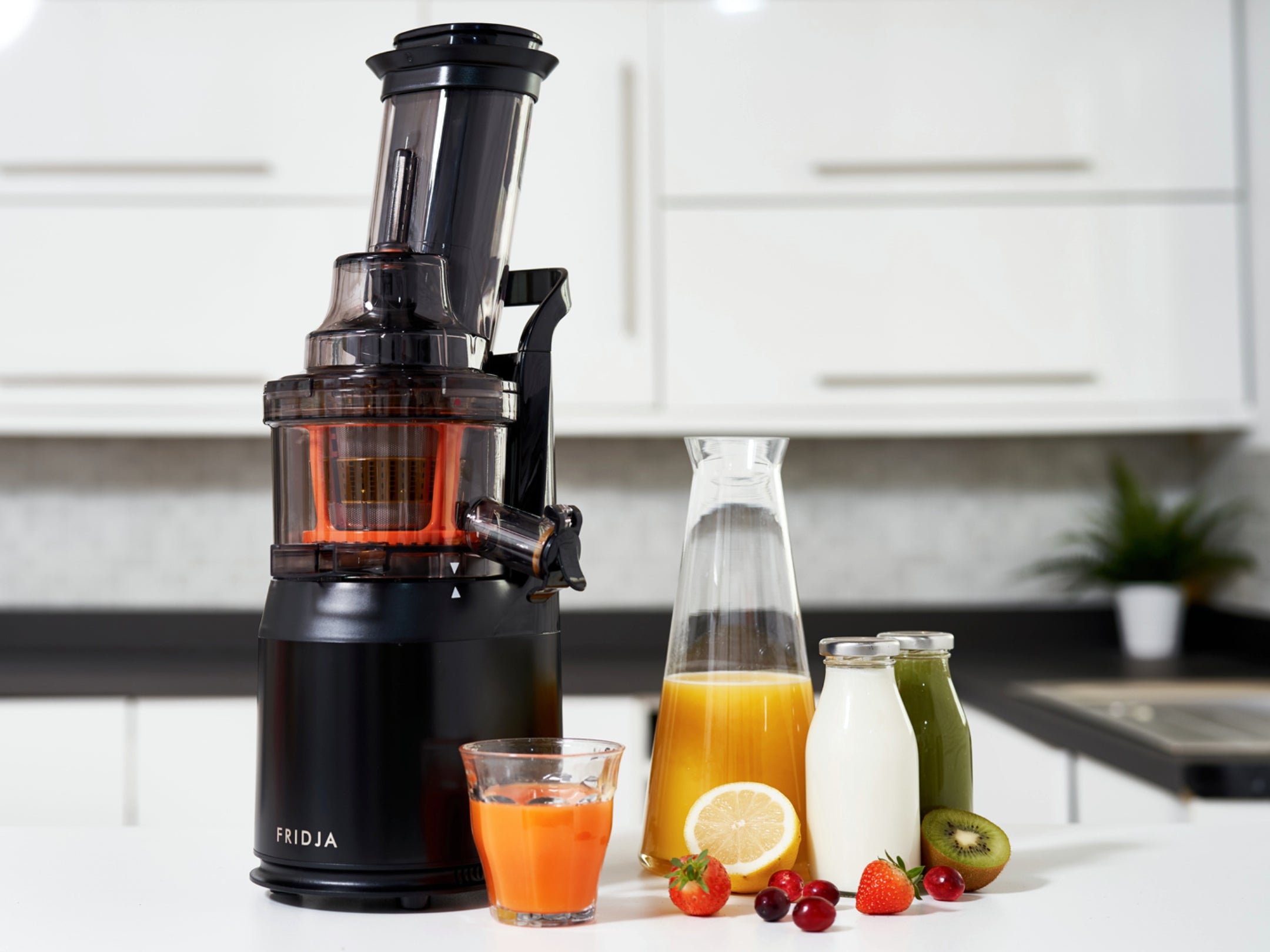 A black juicer is on a kitchen counter with a glass of orange juice, two carafes of juice, and various fruits including lemon, kiwi, strawberry, and cherries arranged beside it.