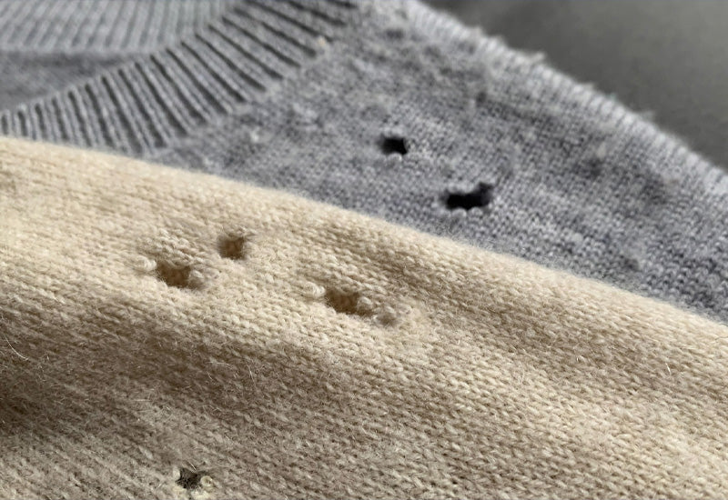 Close-up of two sweaters, one beige and one gray, both with small holes indicating damage, possibly from moths or wear. The fabrics have a soft, woolen texture.