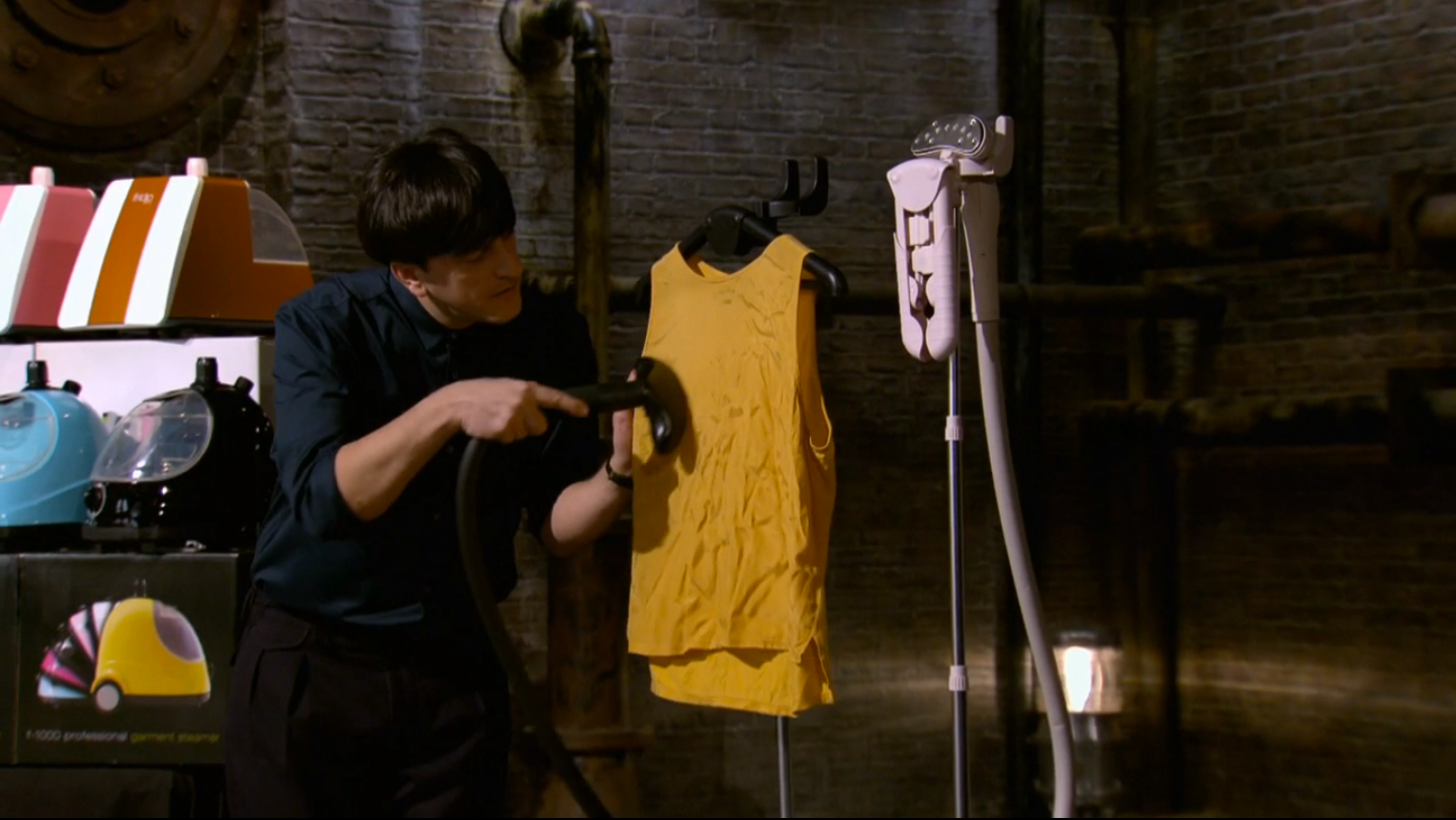 A person uses a handheld steamer to remove wrinkles from a yellow sleeveless shirt hanging on a rack, with other steaming equipment and garments visible nearby in an industrial-style setting.
