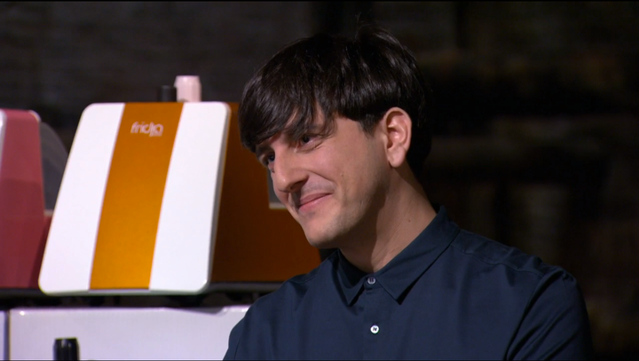 A person with short dark hair, wearing a dark button-up shirt, smiles slightly while looking to the side. Behind them is an orange and white machine labeled fricja.