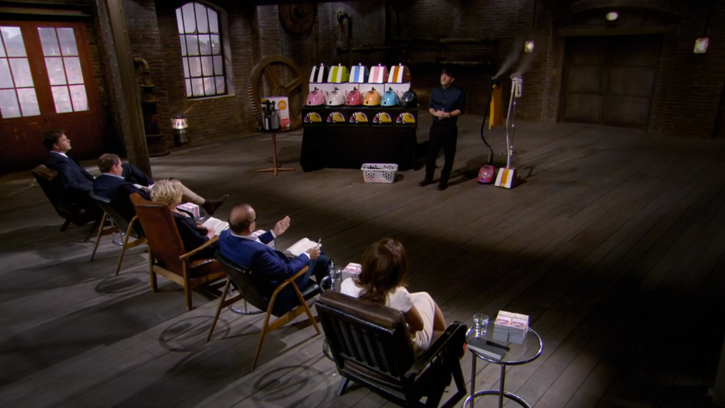 A person presents a product display to four seated judges in a large, industrial-style room with wooden floors and brick walls. Various colorful items are arranged on a table and stand.
