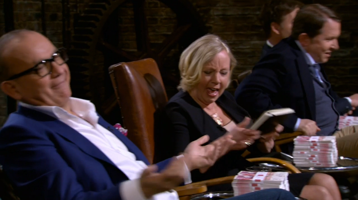 Four people in business attire sit in chairs, reacting energetically. The nearest man gestures with his hands, the woman beside him appears to be speaking or laughing and holding a notebook, and stacks of cash are visible on the table.