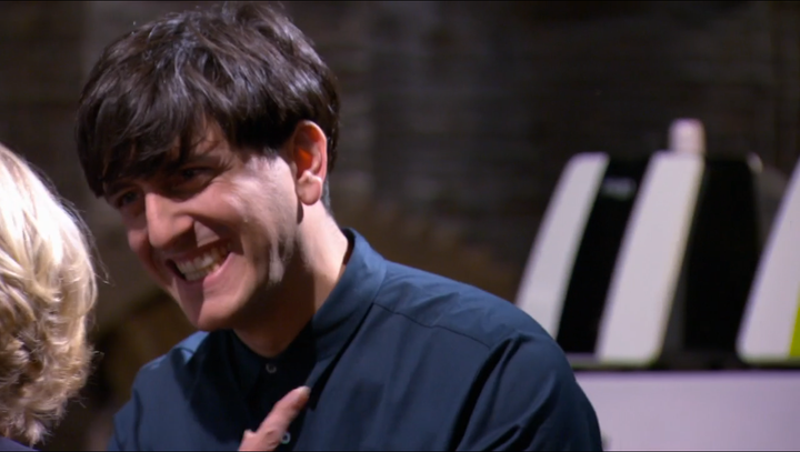 A man with dark hair, wearing a dark shirt, smiles warmly and gestures with his hand near his chest. The background is blurred with some modern equipment visible.