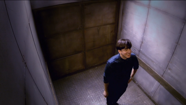 A person with short dark hair, wearing a dark shirt and pants, stands and smiles in the corner of a metallic, dimly lit room with ribbed walls and a large metal door.