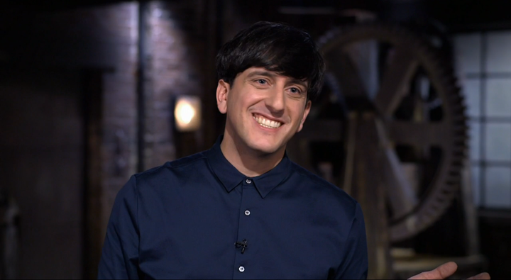 A man with dark hair, wearing a navy blue button-up shirt, smiles warmly while standing indoors. The background is blurred, showing industrial-style decor with brick walls and large gears.