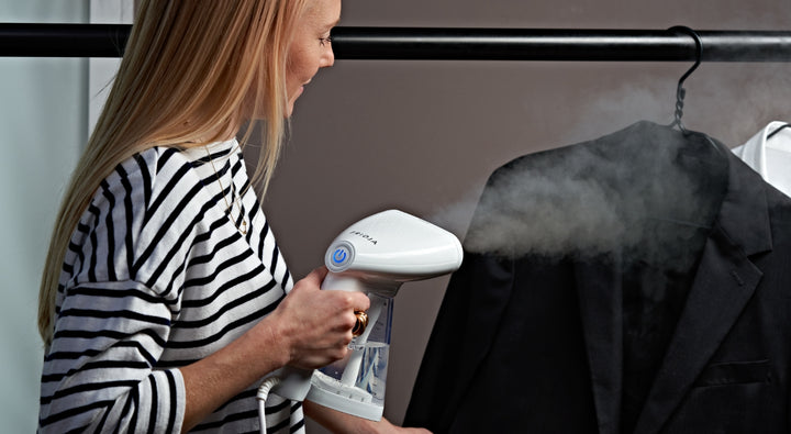 A woman in a striped shirt uses a handheld garment steamer to remove wrinkles from a black suit jacket hanging on a rack, with visible steam coming from the device.