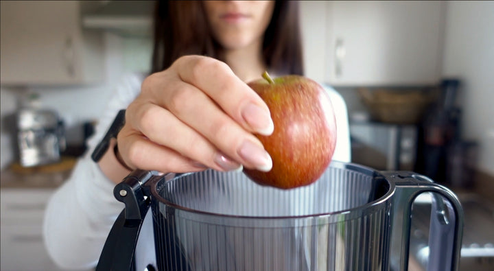 A woman holds a red apple above a blender in a kitchen, preparing to add it. The background is blurred, focusing on her hand, the apple, and the blender.
