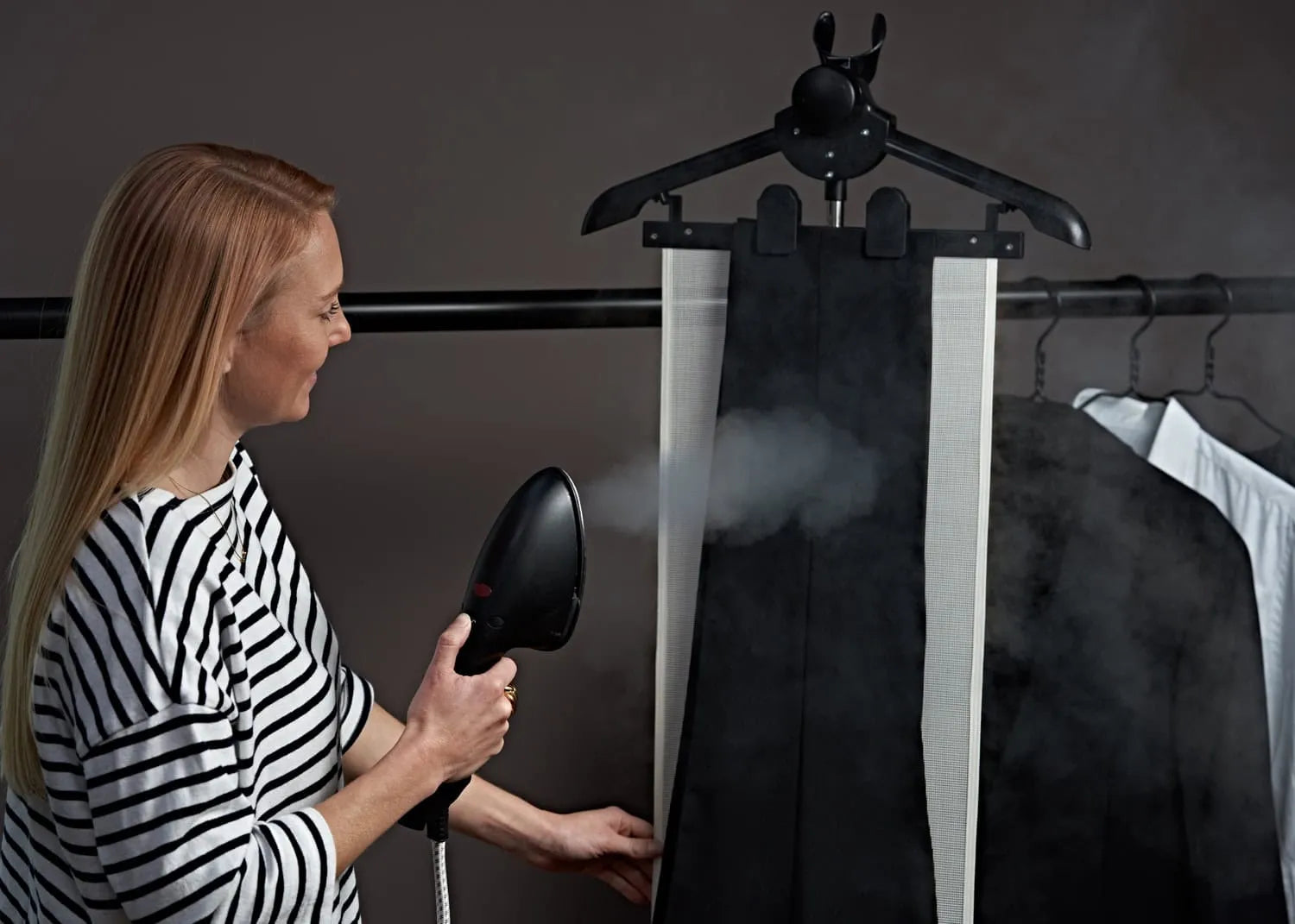 A woman in a striped shirt uses a handheld steamer to remove wrinkles from a pair of black pants hanging on a hanger, with steam visibly rising. Other clothing items hang in the background.