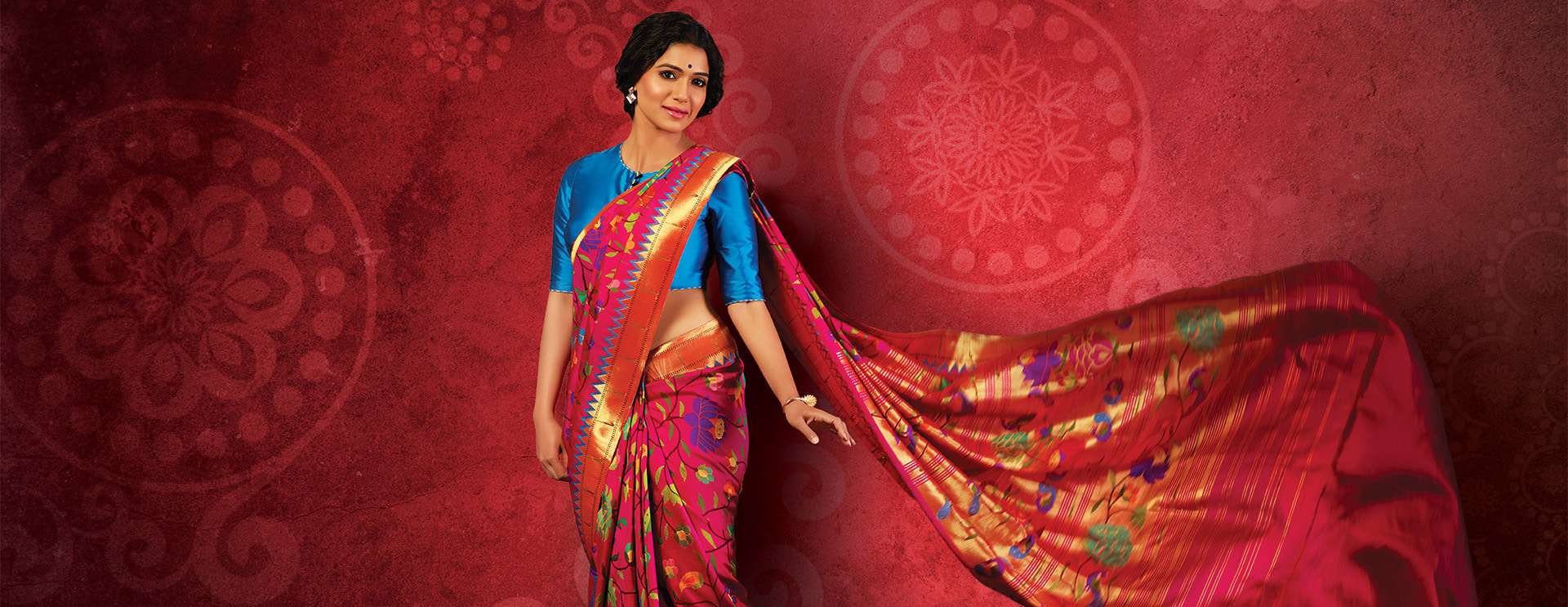 A woman in a vibrant pink and gold saree with a blue blouse poses gracefully against a textured red background with decorative circular patterns. The saree’s pallu flows elegantly behind her.