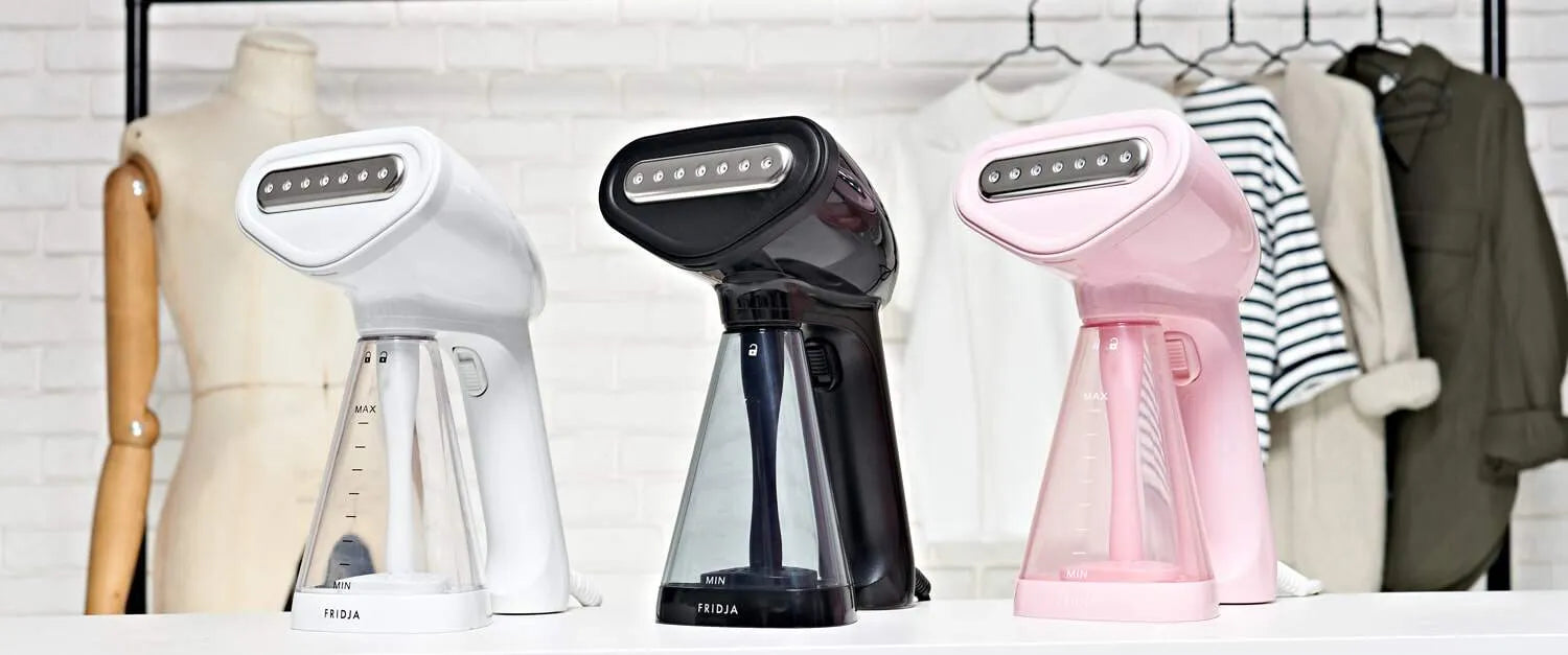 Three handheld garment steamers—white, black, and pink—are displayed on a table in front of a clothing rack with various clothes and a dress form against a white brick wall.