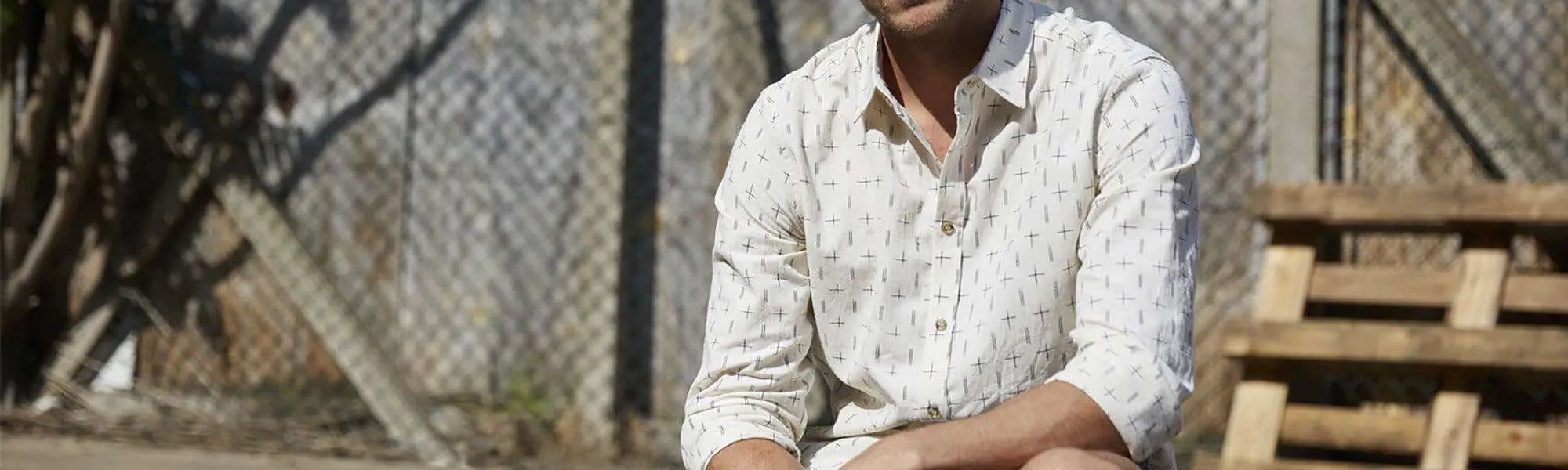 A man in a light-colored, patterned button-up shirt sits outdoors on a wooden pallet, with a chain-link fence and trees in the background.