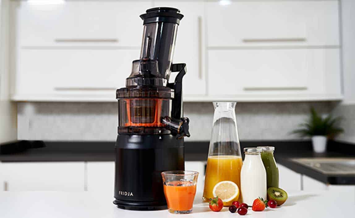 A black juicer sits on a kitchen counter with a glass of orange juice, a carafe of yellow juice, a bottle of green juice, and assorted fruits including strawberries, kiwi, grapes, and lemon in front.