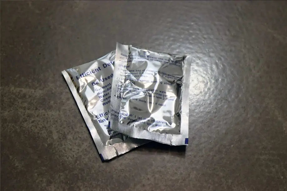 Two small, silver, sealed packets lie overlapping on a textured gray surface, likely containing desiccant or moisture absorber material.