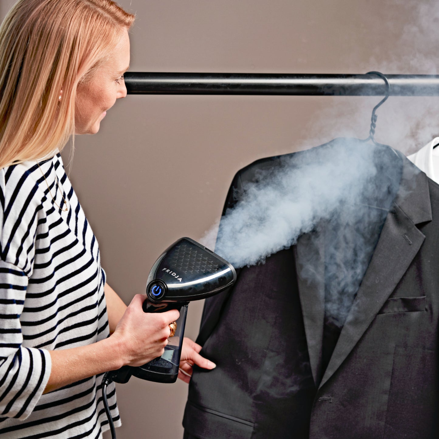 Wearing a striped shirt, a person uses the Fridja f10 Handheld Clothes Steamer - Refurbished Grade A to remove wrinkles from a black suit jacket on a rack, with visible steam highlighting this compact garment care solution.
