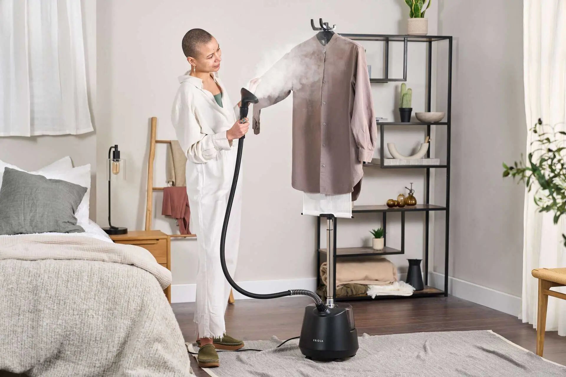 A person uses a clothes steamer to remove wrinkles from a shirt hanging on a stand in a tidy, modern bedroom with neutral decor and shelves in the background.