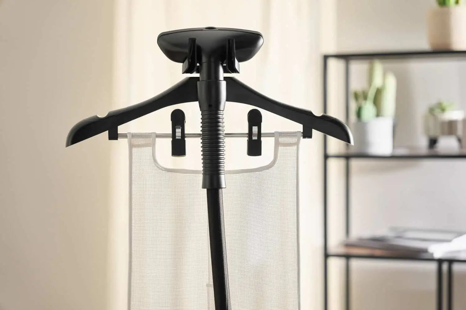 A close-up of a garment steamer with a hanger and clips holding a piece of fabric, set indoors with a blurred background featuring shelves and potted plants.