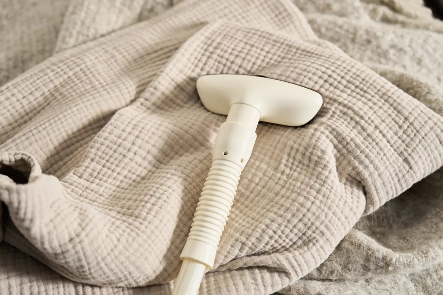A handheld fabric steamer is resting on a light beige, textured blanket with a waffle pattern. The blanket appears soft and slightly wrinkled.
