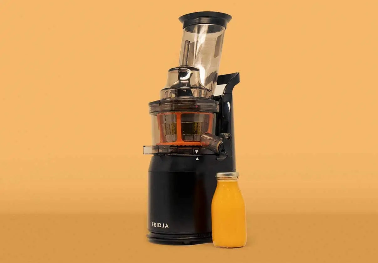 A black Fridja juicer is displayed against a solid tan background, with a small glass bottle filled with orange juice placed beside it.