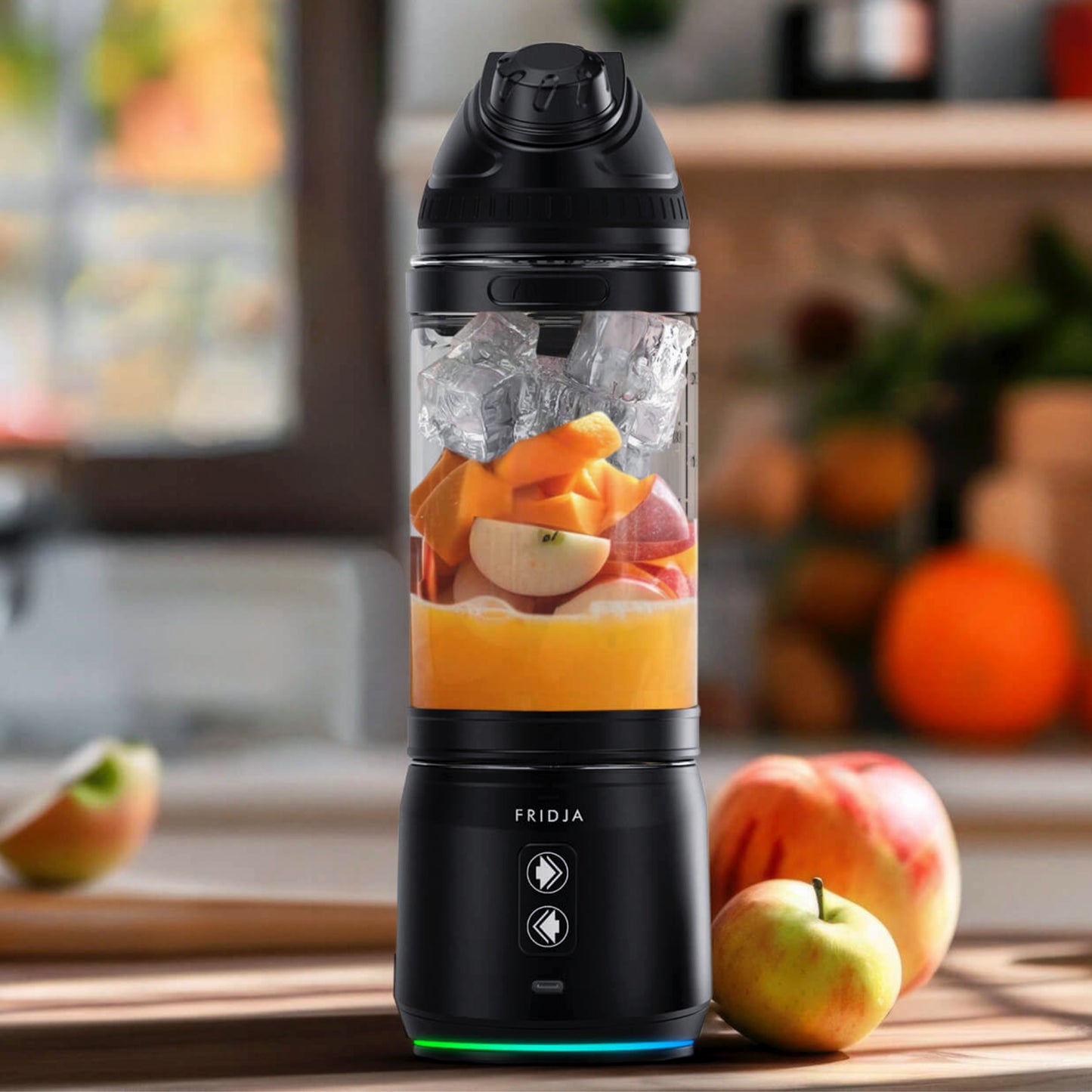 A Fridja f500 Portable Blender in black, filled with ice, chopped fruit, and juice, sits on a kitchen counter with apples and blurred kitchen items in the background.
