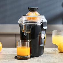 The Fridja f800 Automatic Citrus Juicer in black sits on a counter, dispensing fresh orange juice into a glass, with another full glass and whole oranges nearby—showcasing the power of this juicing machine.