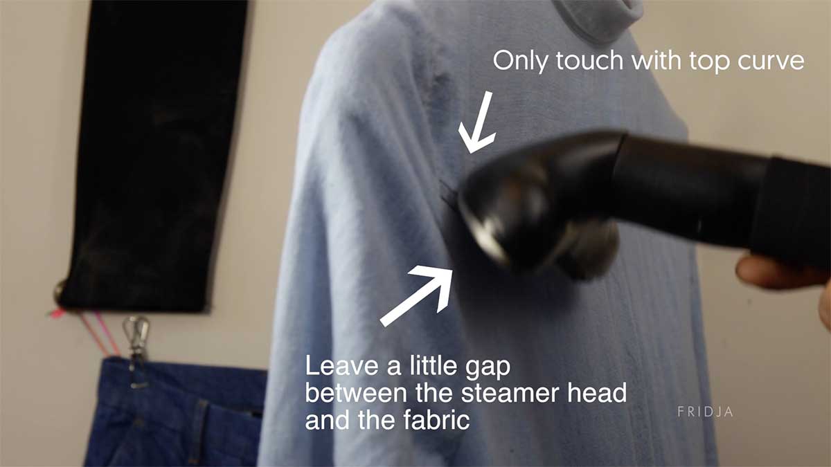 A garment steamer is being used on a light blue shirt. Arrows and text indicate to only touch with top curve and leave a little gap between the steamer head and the fabric.