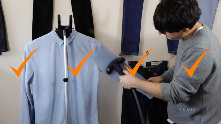 A person uses a garment steamer on a blue button-up shirt hanging on a stand, with orange check marks indicating approval. Pairs of jeans hang in the background.