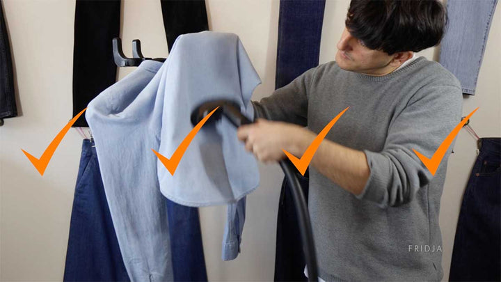 A person uses a garment steamer on a light blue shirt hanging among several pairs of jeans. Four large orange checkmarks are overlaid on the image, indicating approval or correctness.