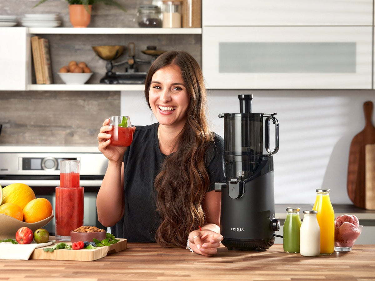 Smiling woman holding a glass of red juice beside the Fridja f2500 Self Feeding Whole Fruit Juicer in a kitchen