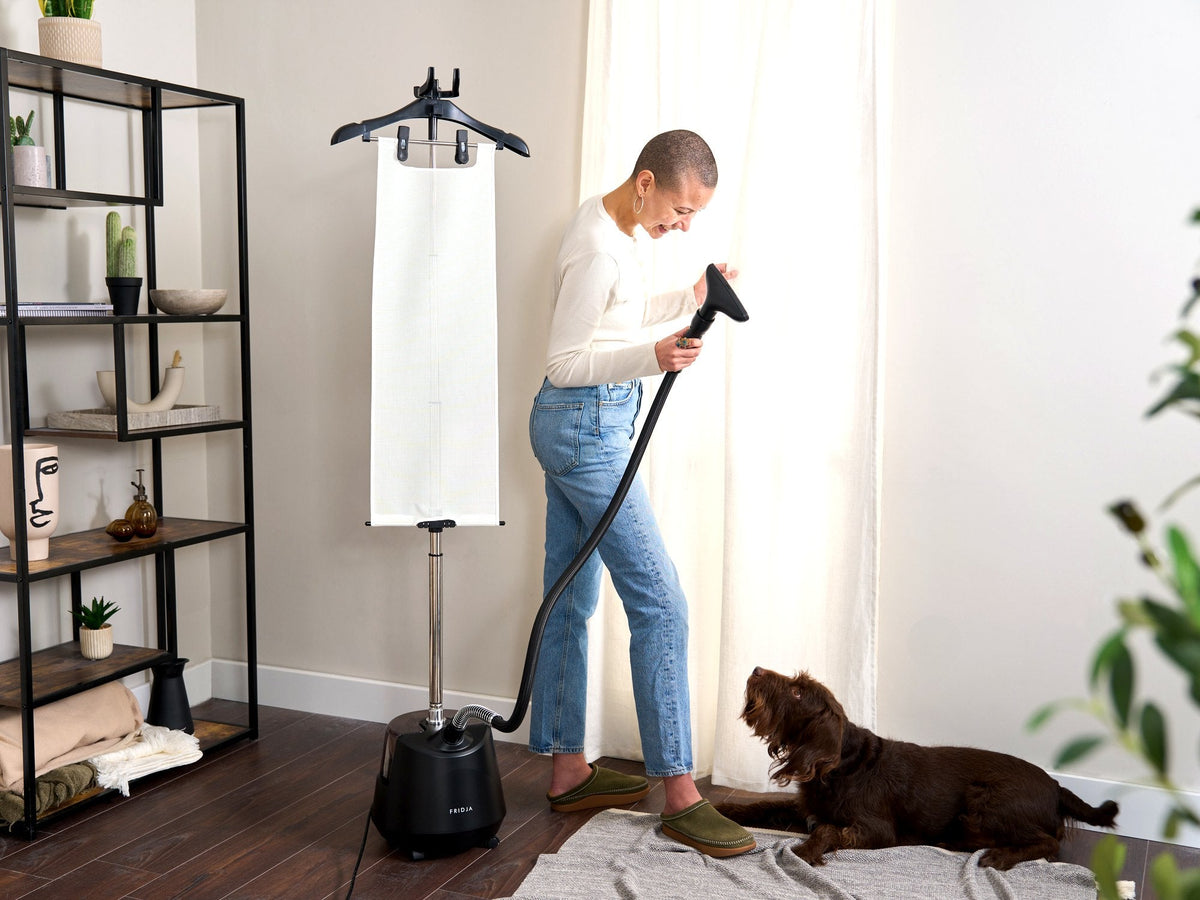 Woman refreshing clothes with the Fridja f1200 Clothes Steamer Pro at home while her dog watches
