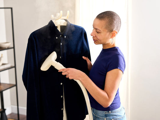 Woman steaming a navy jacket on a hanger with the Fridja f1200 Clothes Steamer Pro in Off White