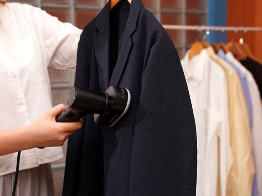 Steaming a navy blazer on a hanger with the Fridja f20 Handheld Clothes Steamer Pro beside a rail of shirts