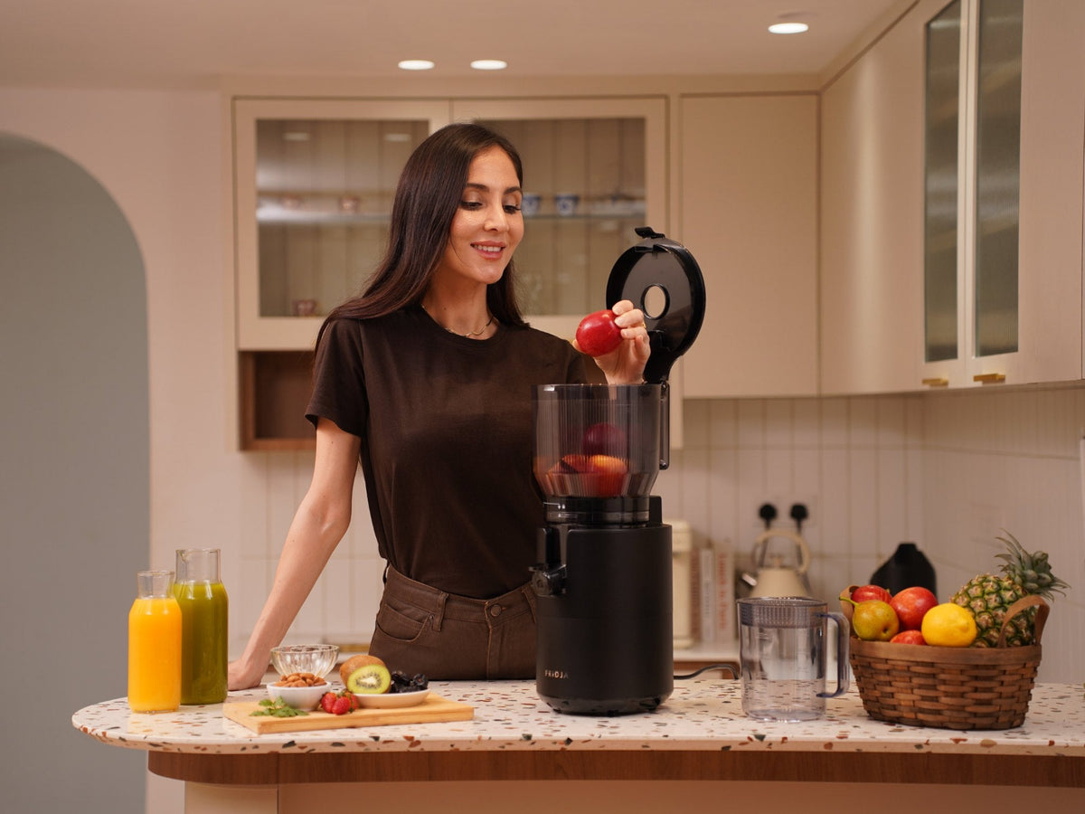 Woman adding an apple to the Fridja f2800 Open Whole Fruit Juicer in a bright kitchen, with fresh juices and a fruit basket on the counter
