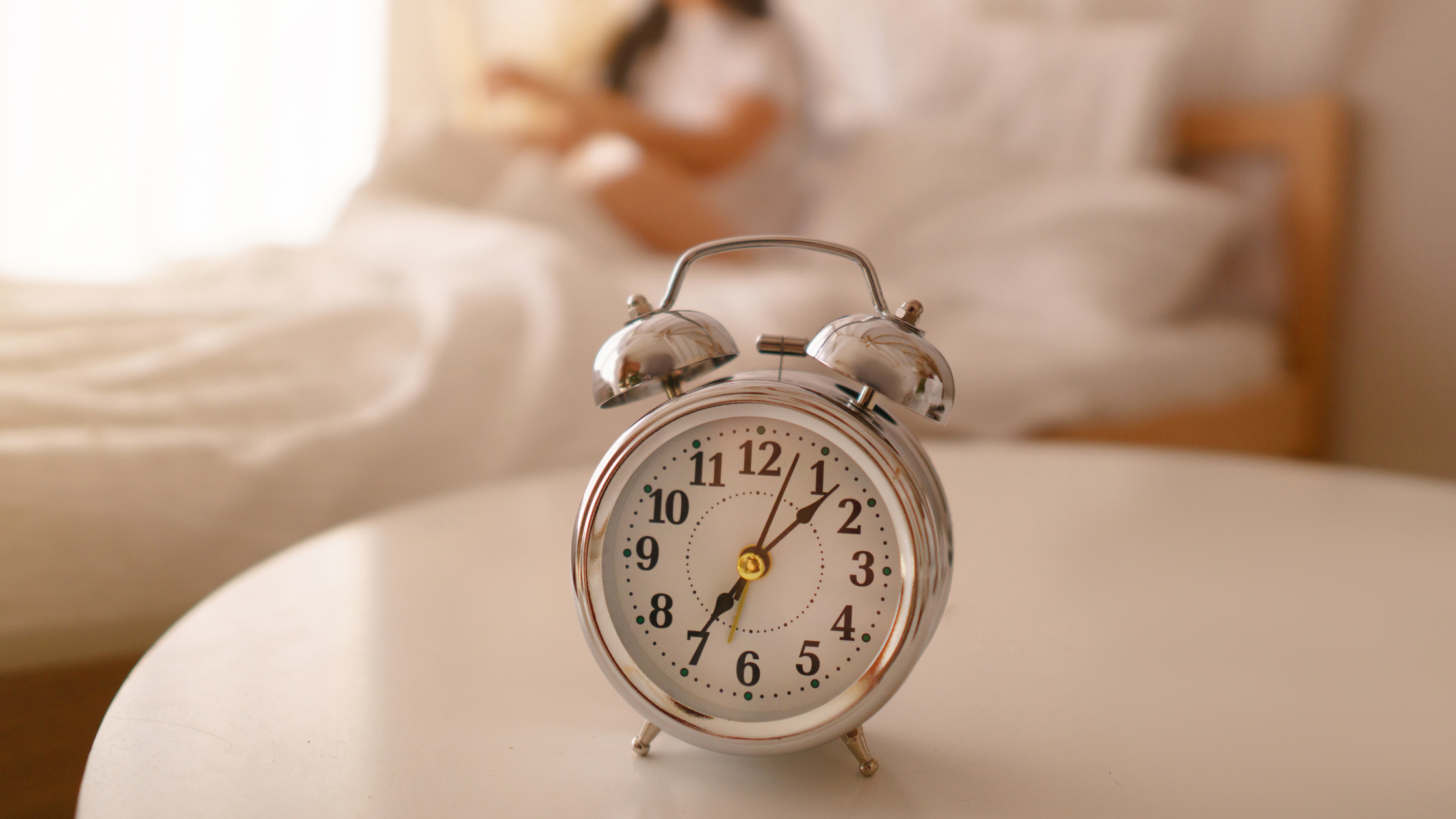 A silver analog alarm clock showing 7:05 sits on a white table in the foreground, with a blurred figure of a person sitting on a bed in a softly lit bedroom in the background.
