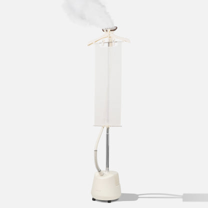 A white vertical clothes steamer with a hanger on top, releasing steam. The device has a water tank at the base, a long pole, and a hose attached to the steaming head, all set against a plain light background.